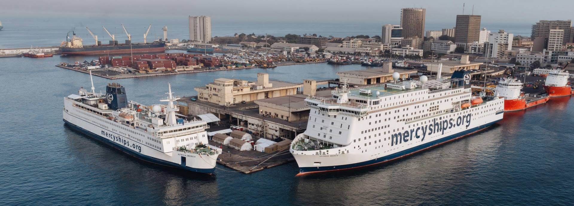 Two hospital ships from the Mercy Ships fleet in port. Mercyships.org painted on the sides.