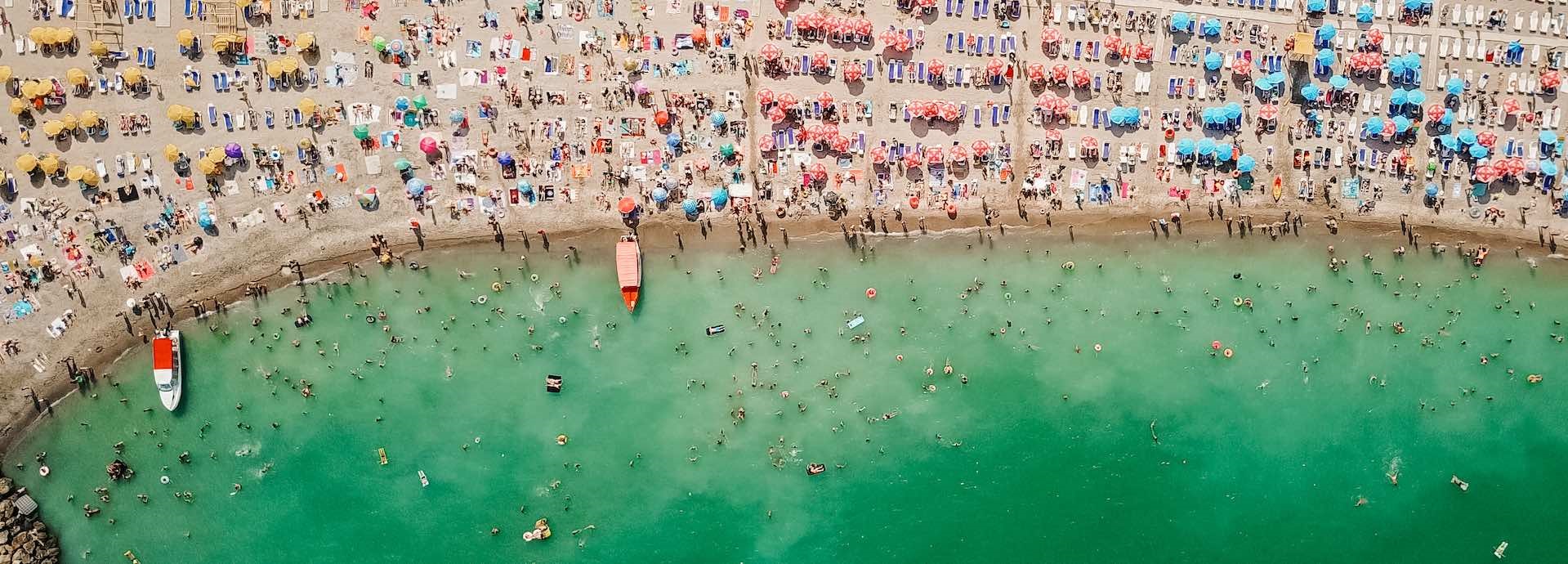 Top-down drone image of a crowded beach filled with umbrellas, towels and shacks. People on land and in the water.
