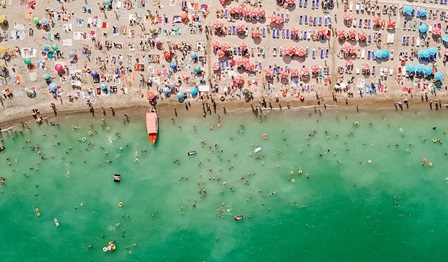 Top-down drone image of a crowded beach filled with umbrellas, towels and shacks. People on land and in the water.