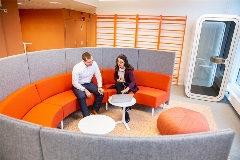 Modern co-working space. Half circle shaped sofa with small round tables in the middle. Two employees chatting in the sofa.