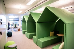 Modern co-working solution: Green cabinets that are shaped like houses. Inside of the cabinets there are sofa-like benches, tables and pillows. One side of the cabin is open.