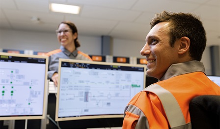 Photo of two smiling employees. One employee closer to the camera facing towards their laptop screen, other one on the other side facing to the camera.