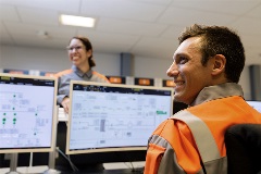 Photo of two smiling employees. One employee closer to the camera facing towards their laptop screen, other one on the other side facing to the camera.